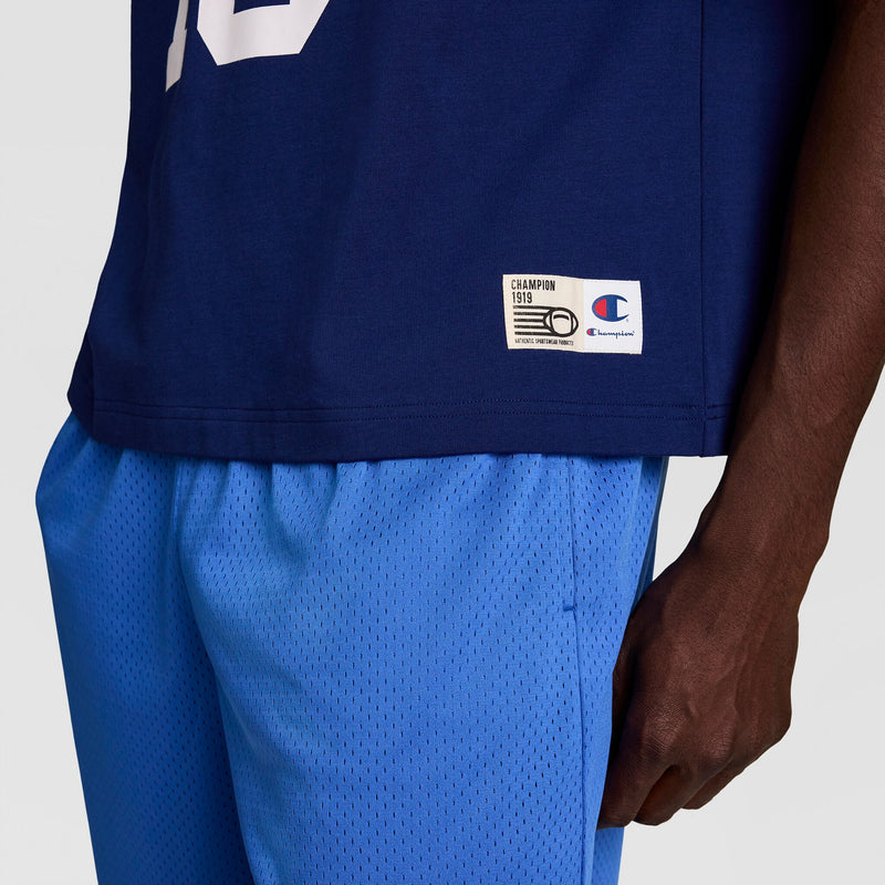 A person wears the Champion Gridiron Football Jersey with a script logo and light blue mesh athletic shorts, shown from the waist down with one arm visible, evoking a vintage athletic style.