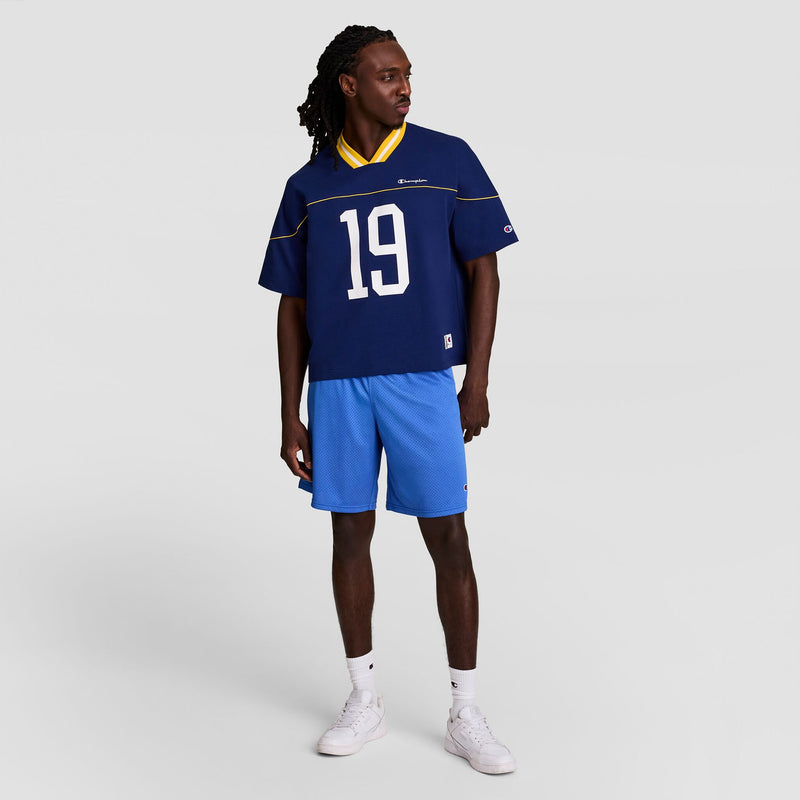 A man with long dreadlocks wears a blue Champion Gridiron Football Jersey with script logo, number 19, light blue shorts, white socks, and white sneakers, standing against a plain light gray background.