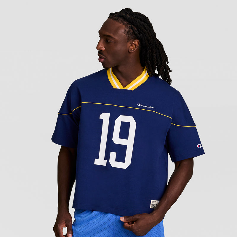 A man with long braids wears the Champion Gridiron Football Jersey, Script Logo, featuring a blue body, yellow-trimmed collar, number 19, and matching blue shorts, standing against a plain light background.