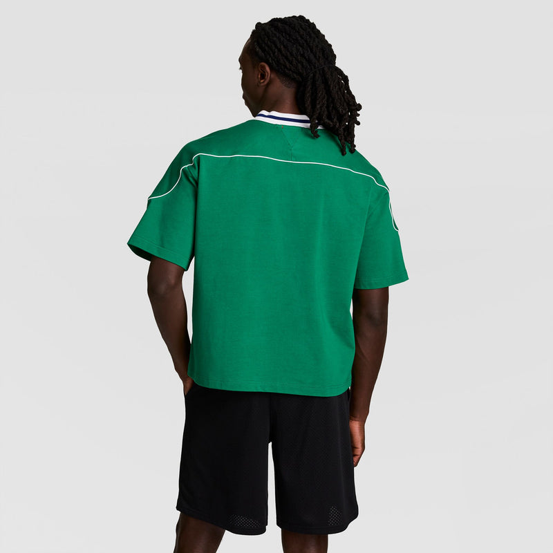 A person with long, braided hair stands against a plain light gray background, wearing the Champion Gridiron Football Jersey, Script Logo in green with white piping and black shorts.