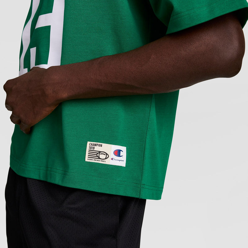 A person is shown from the torso down wearing a green Champion Gridiron Football Jersey with script logo and black shorts. Their left hand rests on their waist, with the Champion brand label visible on the jersey.