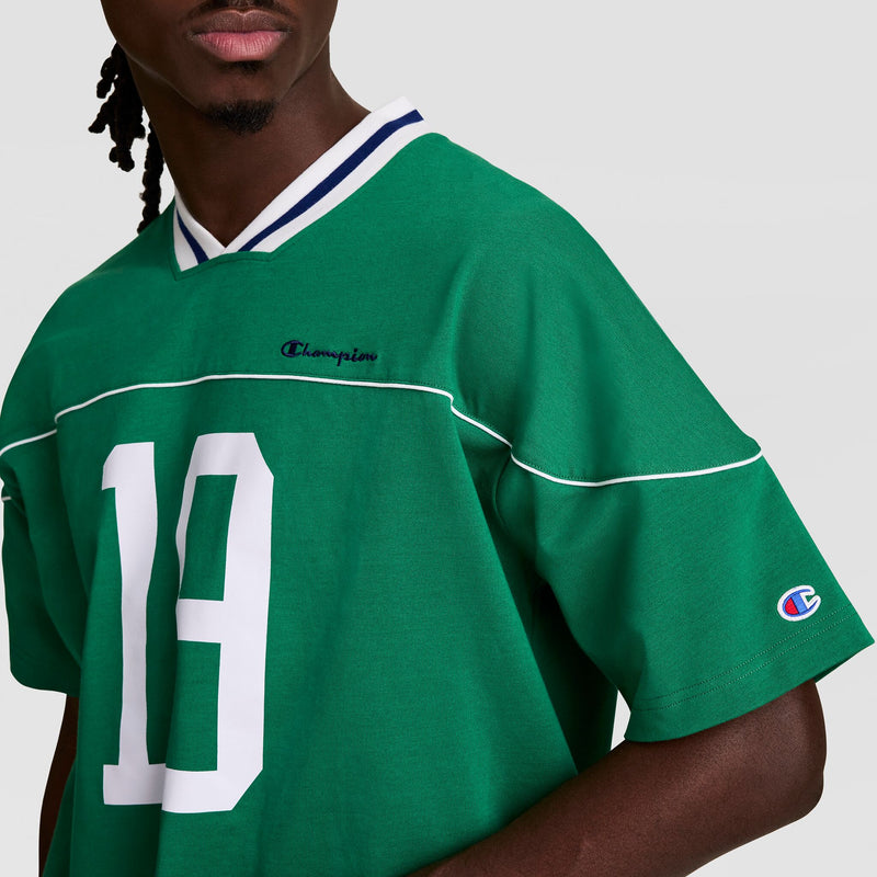 A person wearing the Champion Gridiron Football Jersey with a script logo, green color, white number 19, vintage athletic white piping, and a striped V-neck collar is seen from the shoulders up against a plain background.