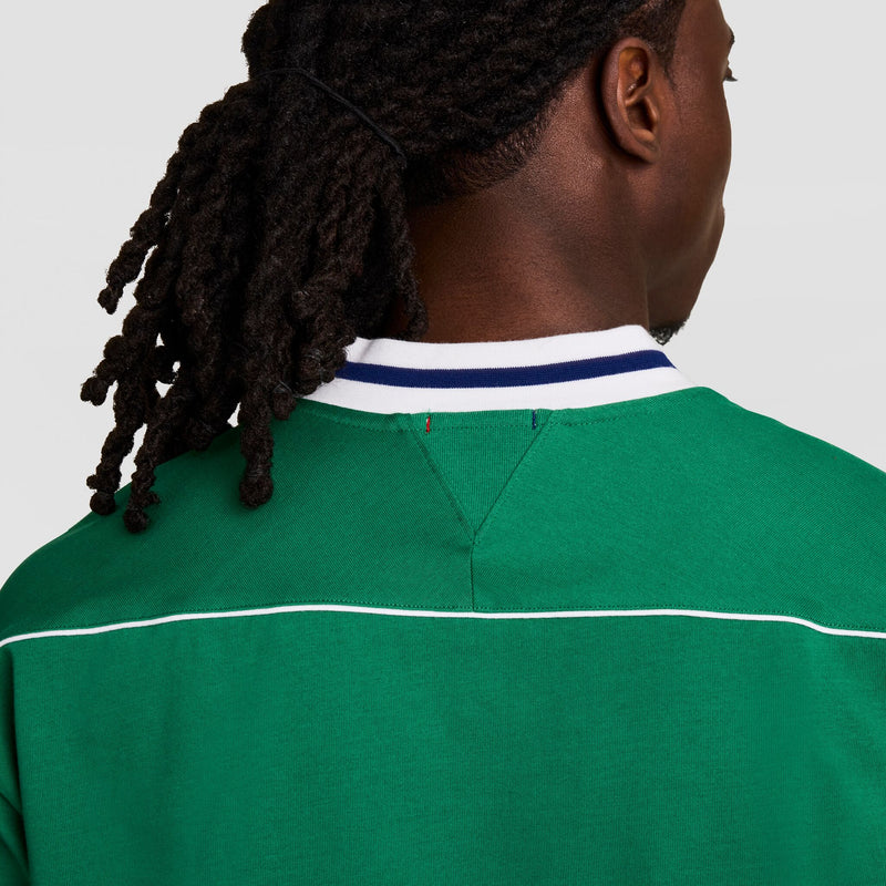 A person with long, dark dreadlocks is shown from behind, wearing the Champion Gridiron Football Jersey, Script Logo—a vintage-style green shirt with white piping and a collar featuring blue and white stripes.