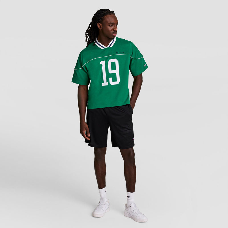 A man with long hair stands against a plain background wearing Champion's Gridiron Football Jersey, Script Logo in green (#19), black shorts, white socks, and white sneakers. He looks left with one hand in his pocket.