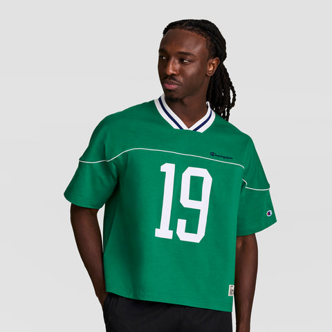 A man with long braids wears the Champion Gridiron Football Jersey, Script Logo in green with a white number 19 and striped collar, channeling vintage athletic style as he looks to his left against a plain light background.