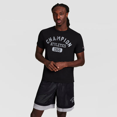 A man stands against a plain background, wearing a black Champion Gridiron Reversible T-Shirt with C Logo and black athletic shorts with white trim, looking at the camera with his hands loosely clasped.