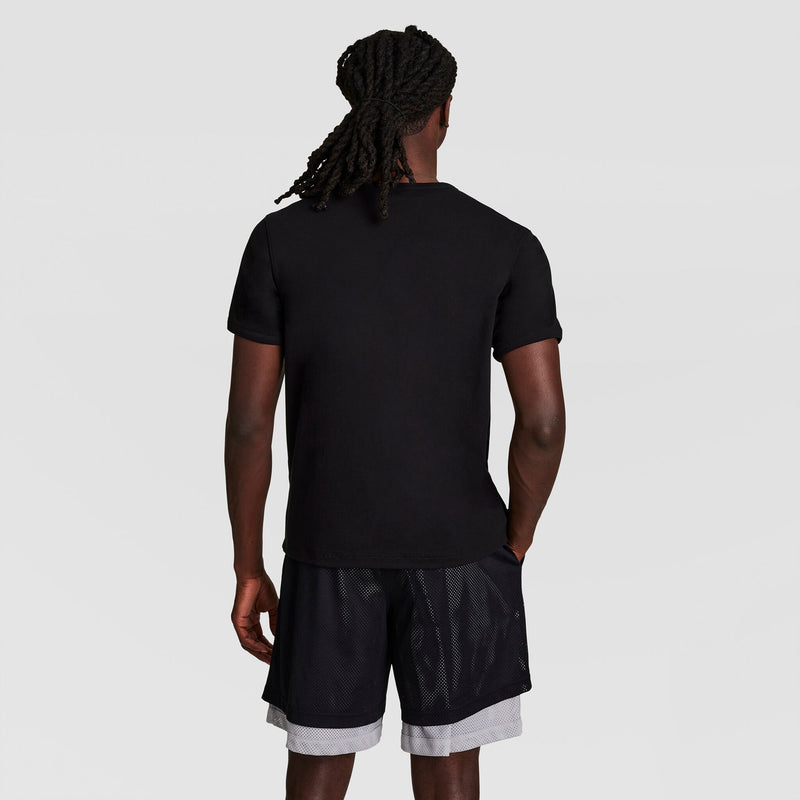 A person with long, braided hair stands with their back to the camera, wearing a black Champion Gridiron Reversible T-Shirt, C Logo and black shorts with white lining against a plain light background.