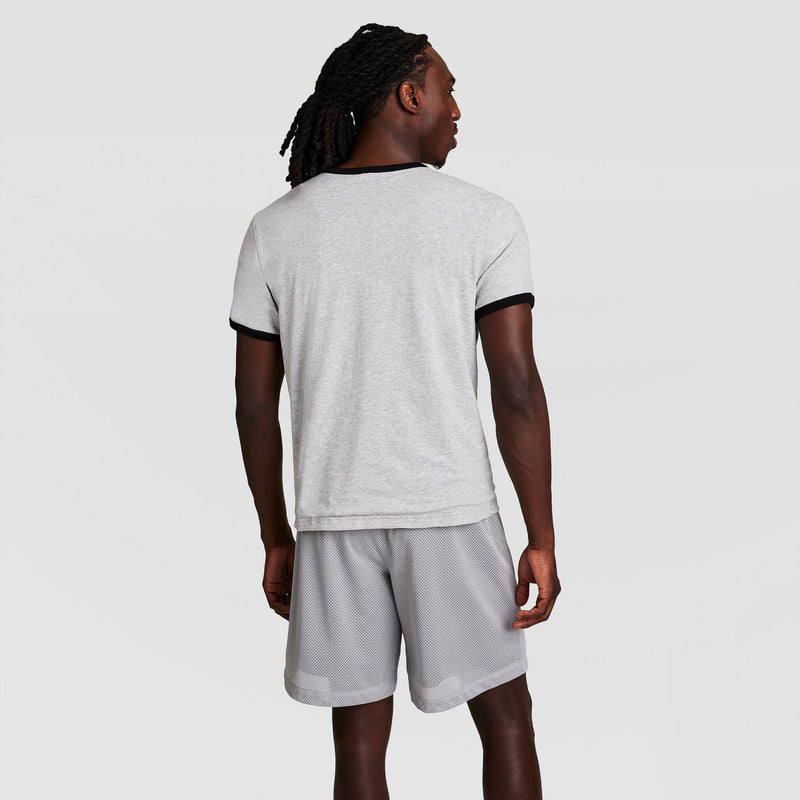A man with long, braided hair faces away, wearing a light gray Champion Gridiron Reversible T-Shirt with C Logo and matching gray athletic shorts, set against a plain white background.