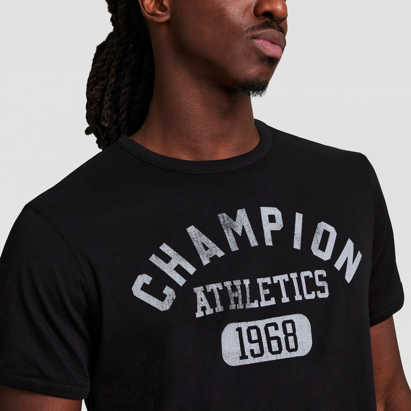 A person in a black Gridiron Reversible T-Shirt by Champion, featuring the C Logo, with “CHAMPION ATHLETICS 1968” in white on the front. The face is partially out of frame.