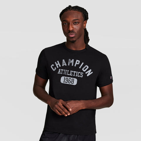 A man with long braided hair wears a black Champion Gridiron Reversible T-Shirt, C Logo. He stands facing forward against a plain, light background, hands loosely clasped.