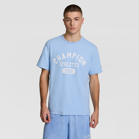 A young man wearing a light blue Champion Athletics 1968 t-shirt and matching athletic shorts stands against a plain white background, looking at the camera with a neutral expression.
