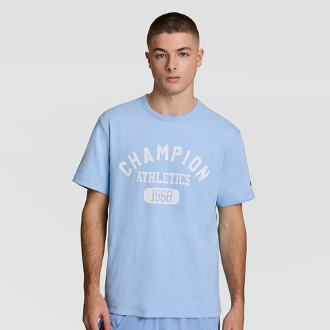 A young man with short hair wears a light blue Champion Gridiron Reversible T-Shirt featuring the C logo. He stands facing forward against a plain, light background.