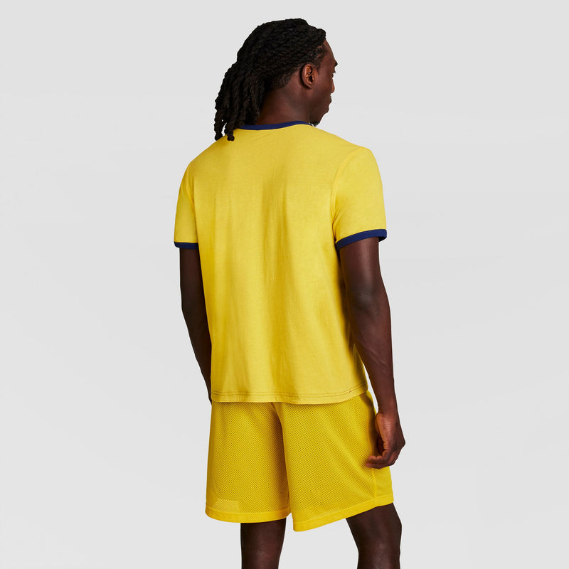 A person with dark, braided hair faces away, wearing a yellow Gridiron Reversible T-Shirt, C Logo by Champion and matching yellow shorts against a plain light gray background.