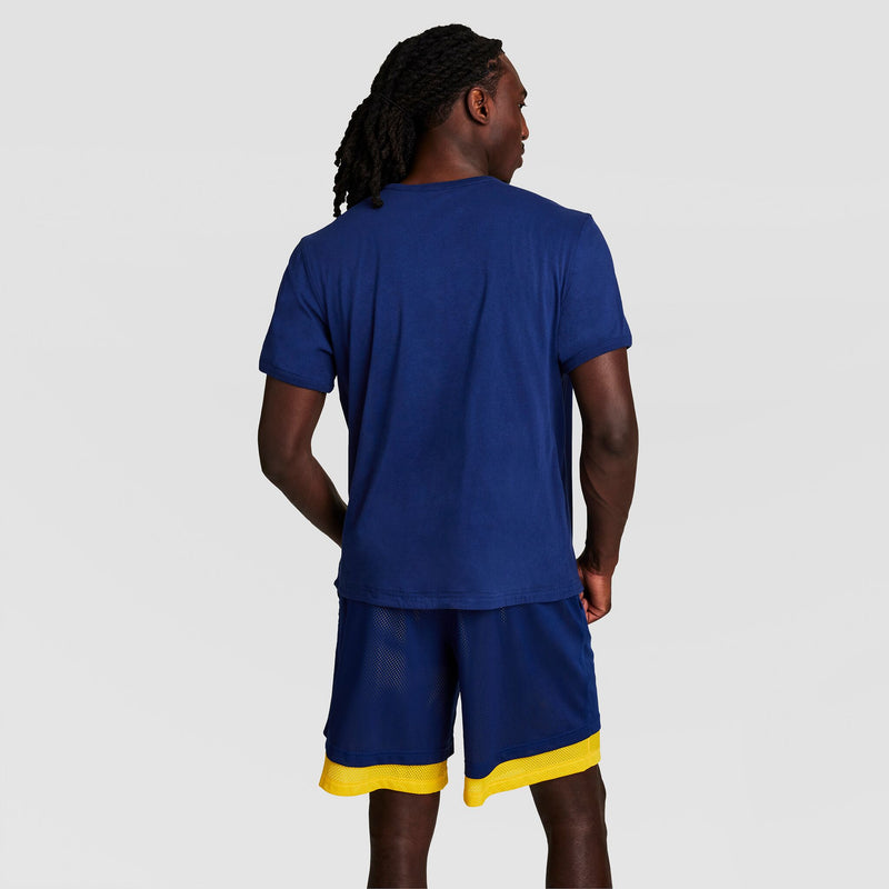 A person with long, braided hair stands with their back to the camera, wearing a navy blue Champion Gridiron Reversible T-Shirt (C Logo) and navy blue athletic shorts with yellow trim against a light gray background.
