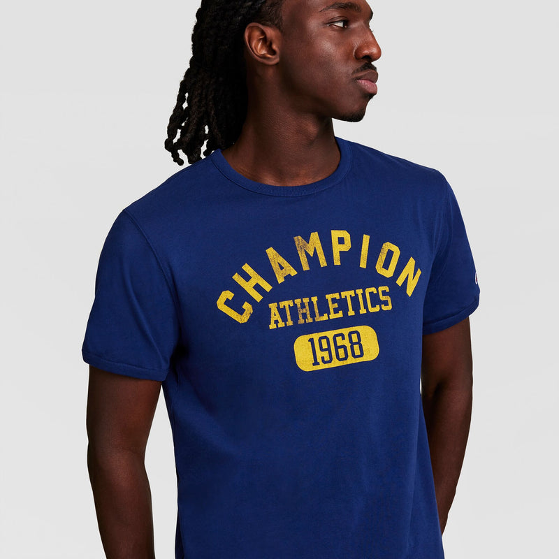 A man in a blue Champion Gridiron Reversible T-Shirt, C Logo, with yellow text stands against a plain light background, looking to his left.