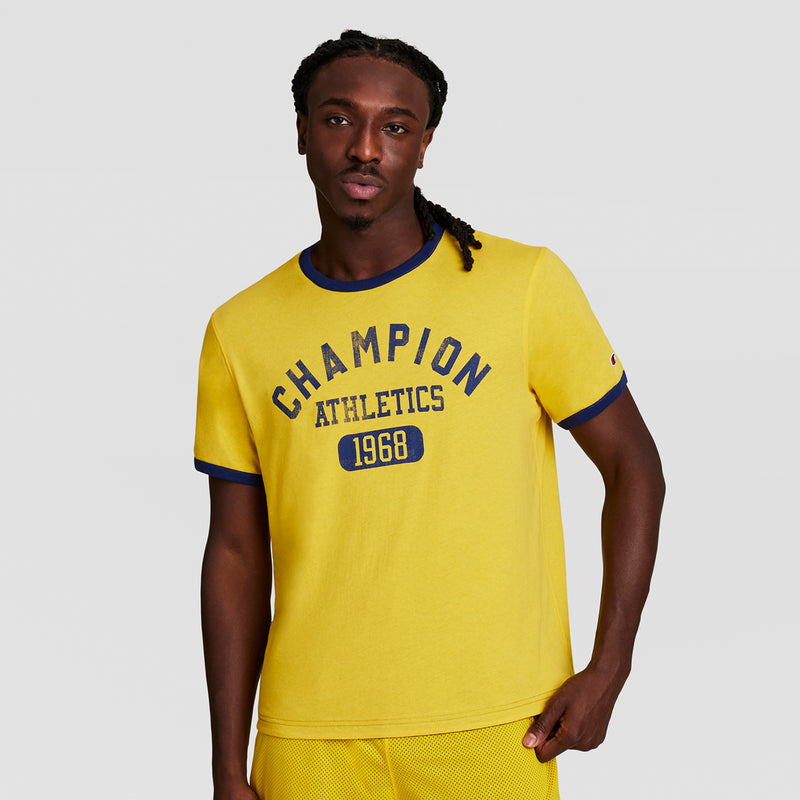 A man with long braids, one hand in his pocket, wears a Champion Gridiron Reversible T-Shirt, C Logo—yellow with blue trim and "CHAMPION ATHLETICS 1968" on the front—standing against a plain light background.
