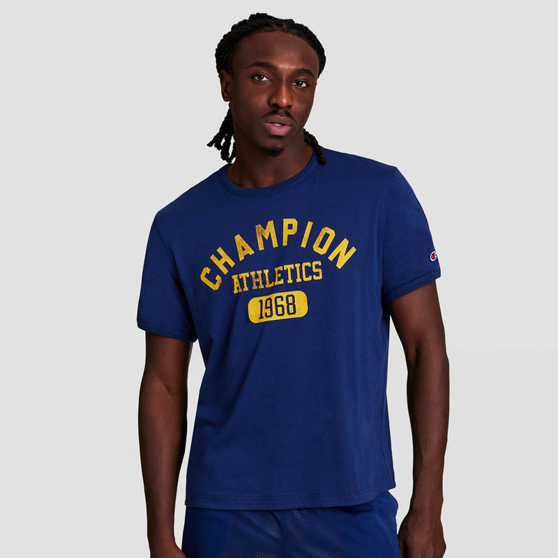 A man wears the Champion Gridiron Reversible T-Shirt, C Logo, featuring blue fabric with yellow “CHAMPION ATHLETICS 1968” text, standing against a plain light gray background.