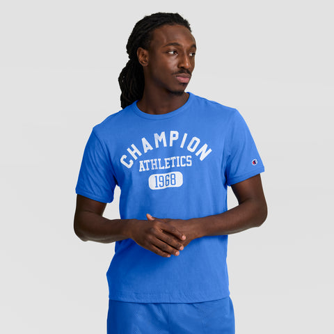 A man wears a blue Champion Gridiron Reversible T-Shirt, C Logo, with matching shorts, standing against a neutral background and looking right with his hands clasped.