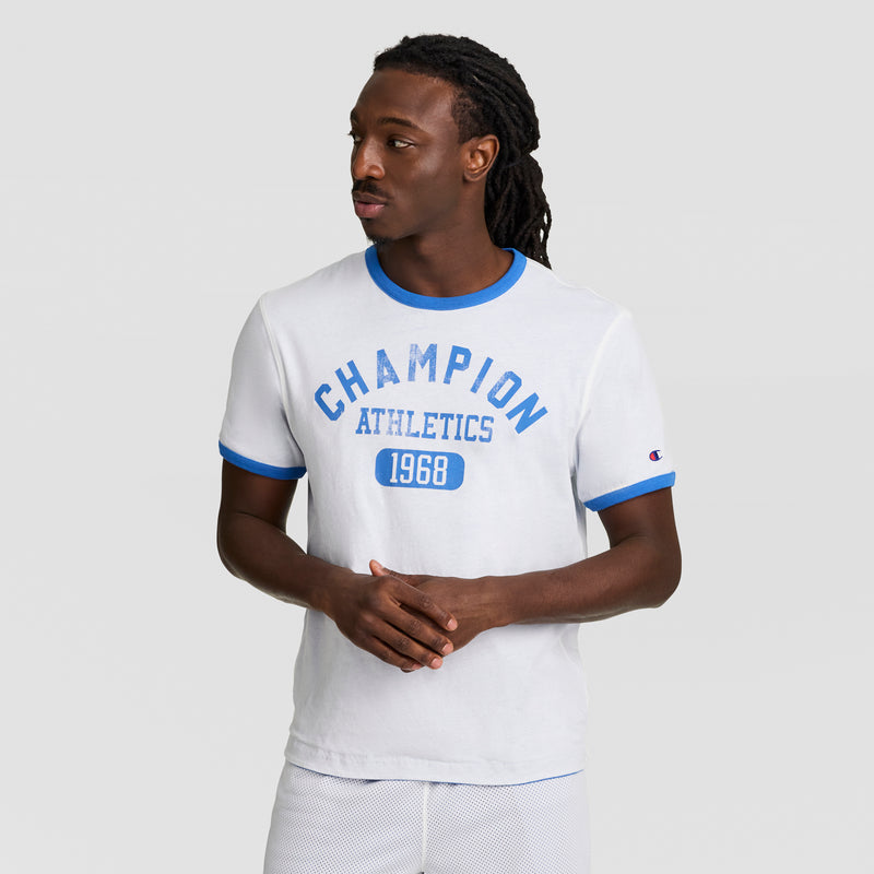 A man with long twisted hair wears a white Champion Gridiron Reversible T-Shirt, C Logo, with blue trim, standing against a plain light gray background and gazing to the side.