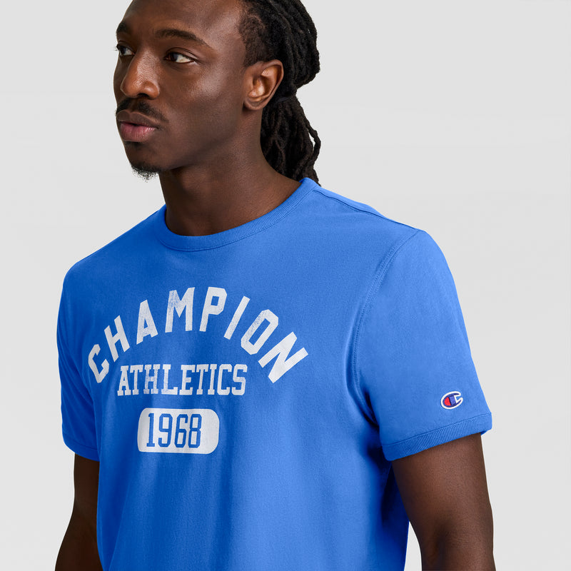 A man with long dreadlocks wears a bright blue Champion Gridiron Reversible T-Shirt, C Logo, featuring “CHAMPION ATHLETICS 1968” in white and a small Champion logo on the sleeve, standing against a plain light background.