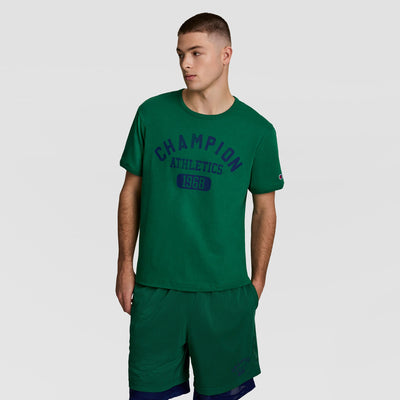 A young man with short brown hair stands against a plain background, wearing a green Champion Gridiron Reversible T-Shirt with the C Logo and matching green shorts, his hands in his pockets.