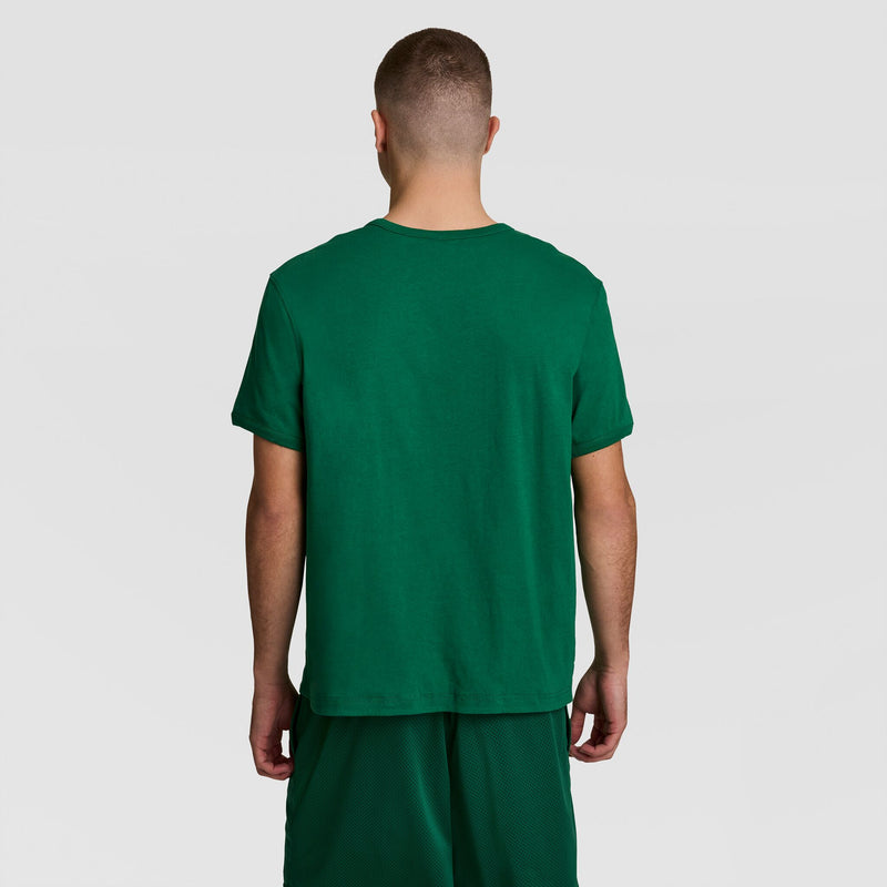 A person stands with their back to the camera, wearing a Champion Gridiron Reversible T-Shirt with C Logo and green shorts against a plain light background.