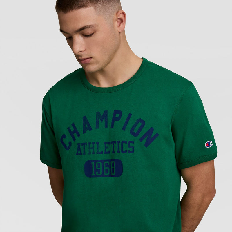 A young man wears the Champion Gridiron Reversible T-Shirt in green, featuring a C Logo on the sleeve, standing against a plain light grey background.