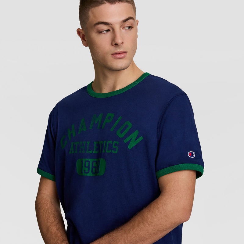 A young man with short hair, looking left against a plain light background, wears a Champion Gridiron Reversible T-Shirt featuring the C Logo.