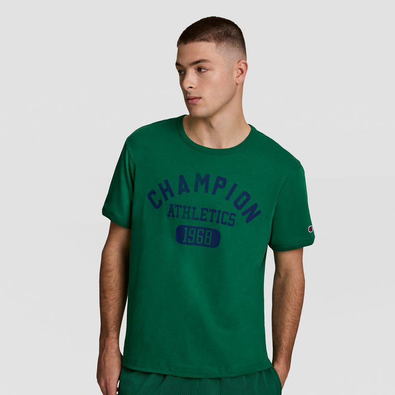 A young man in a green Champion Gridiron Reversible T-Shirt with C Logo and matching shorts stands against a plain light gray background, looking left with a relaxed expression.