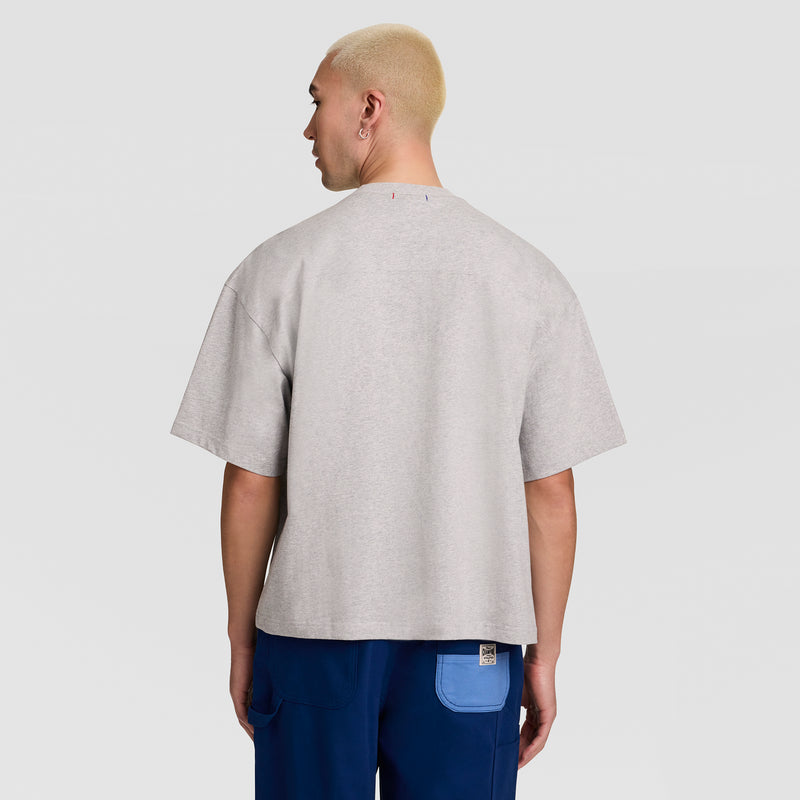 A person with short blond hair wearing a Champion Short Sleeve Boxy T-Shirt, Embroidered Script Logo and blue pants stands facing away against a plain light gray background.