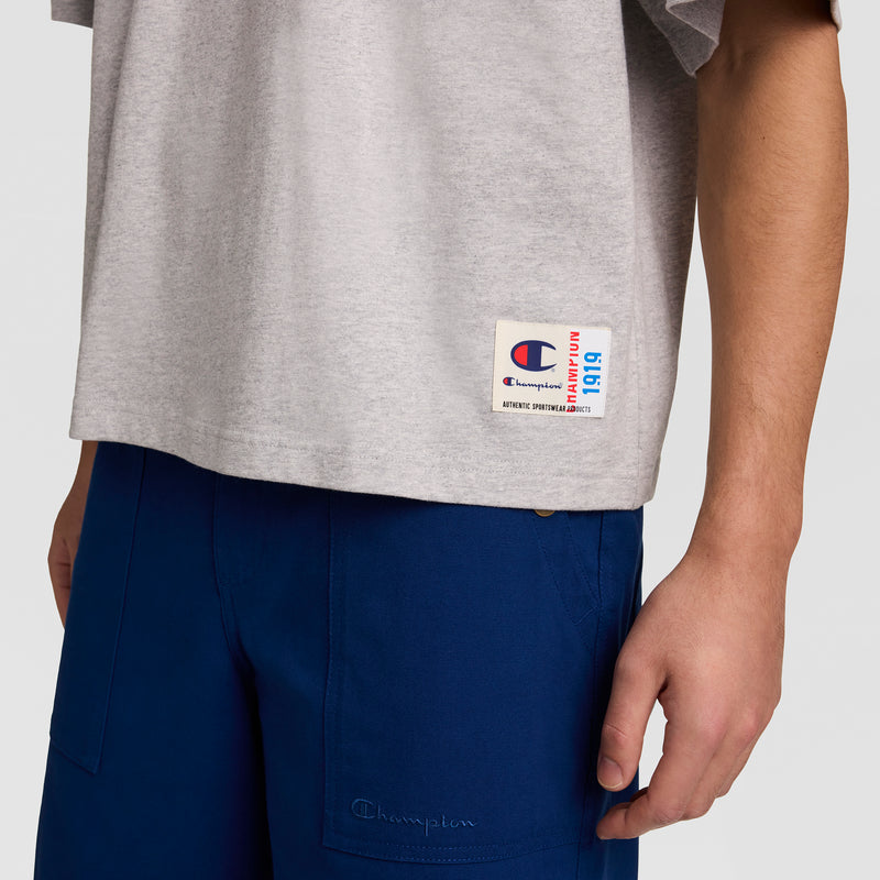 A person wears a gray Champion Short Sleeve Boxy T-Shirt, Embroidered Script Logo, with a logo patch near the hem and blue pants. Only their torso and right arm appear against a plain background.