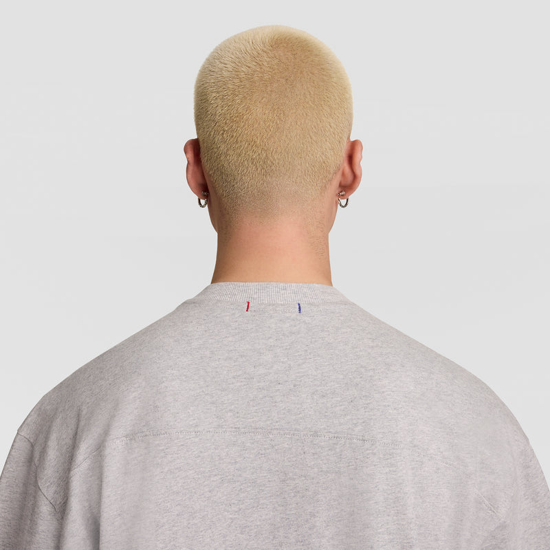 Person with short, bleached blond hair and earrings is seen from the back wearing a light gray Champion Short Sleeve Boxy T-Shirt, Embroidered Script Logo. The background is plain white.