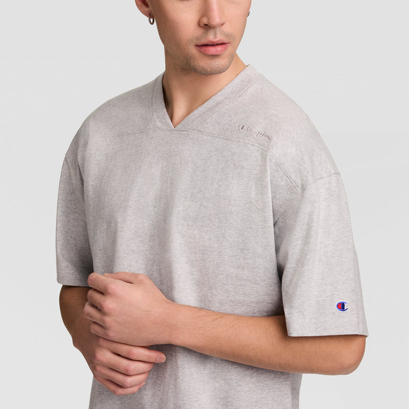 A person in a light gray Champion Short Sleeve Boxy T-Shirt, Embroidered Script Logo, stands with one hand holding their arm, gazing slightly to the side.
