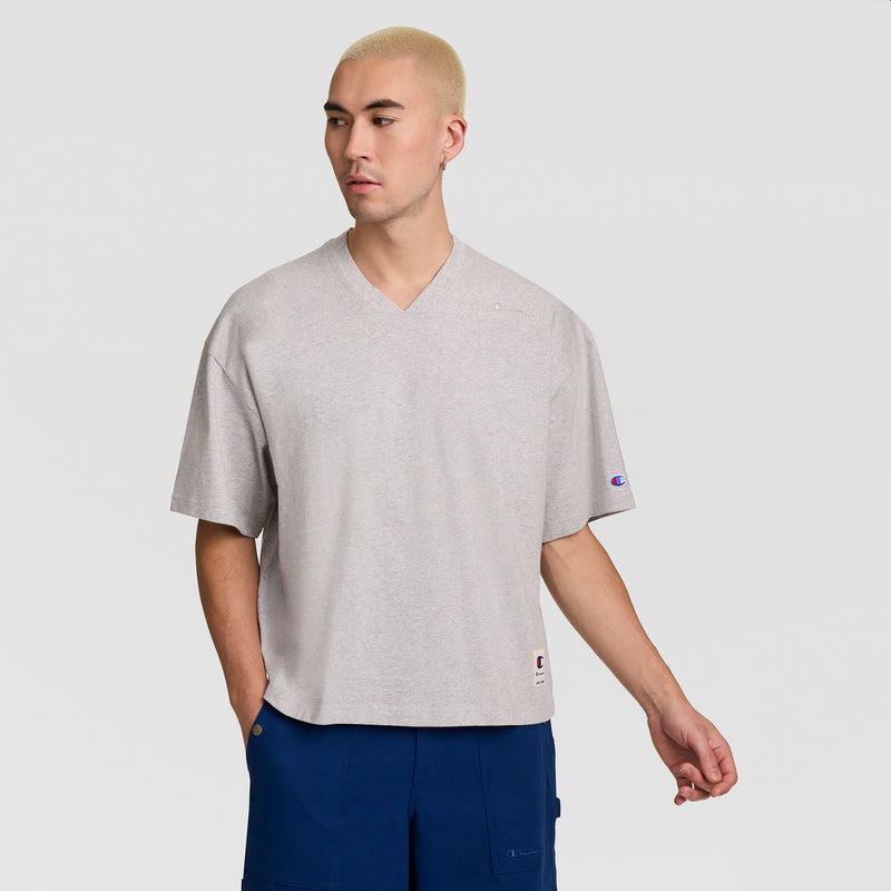 A man with short blond hair wears the Champion Short Sleeve Boxy T-Shirt, Embroidered Script Logo and blue cargo shorts, standing against a light gray background with one hand in his pocket.