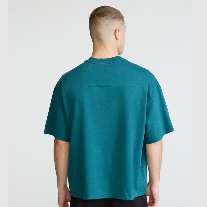 A person with short hair is standing with their back to the camera, wearing the Champion Short Sleeve Boxy T-Shirt with an embroidered script logo in teal and black shorts, against a plain, light background.