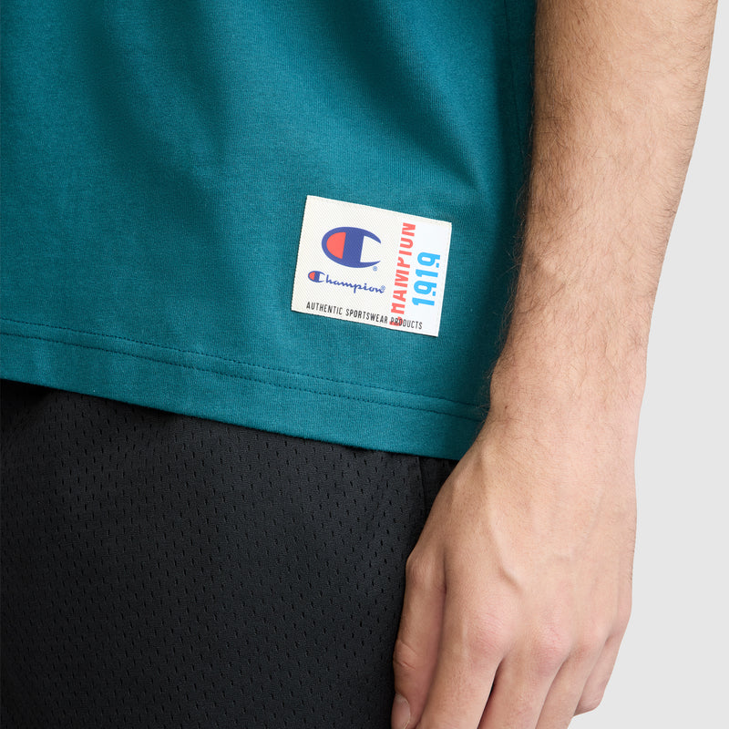 A close-up of a person’s right hand resting by their side, wearing the Champion Short Sleeve Boxy T-Shirt with an embroidered script logo—crafted from soft ring-spun cotton—paired with black mesh shorts and featuring a visible logo tag.