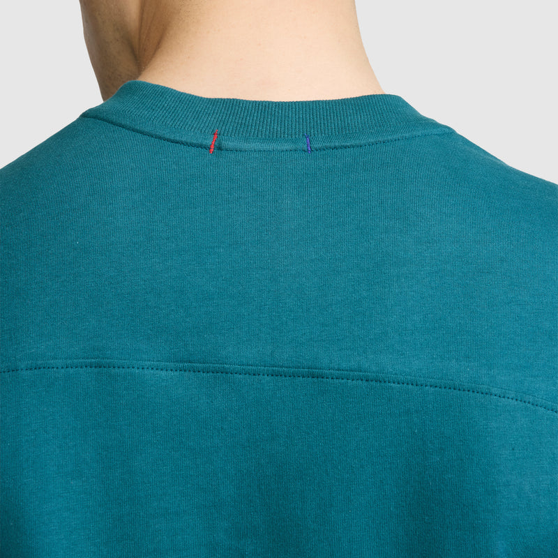 Close-up of the back neckline of a teal Champion Short Sleeve Boxy T-Shirt with an embroidered script logo, crafted from soft ring-spun cotton. The collar features red and blue stitches, and the image is cropped to show only the neck and upper back of a person.