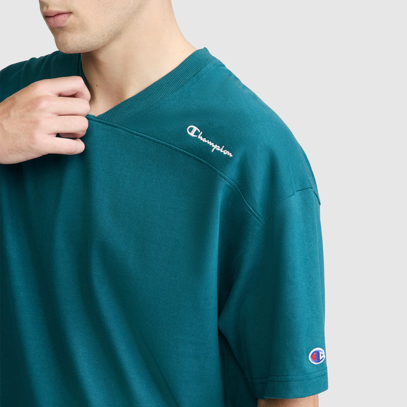 A person wearing a teal Champion Short Sleeve Boxy T-Shirt with an embroidered script logo gently holds the neckline. The Champion branding is visible on the chest and sleeve of this relaxed fit tee. The background is plain and light-colored.