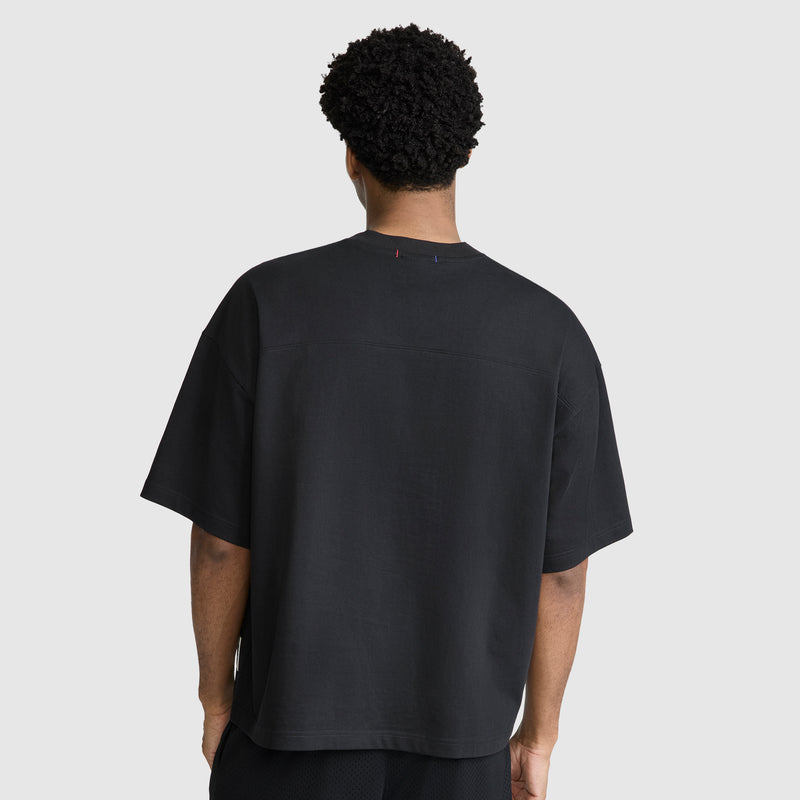 A person with short curly hair is standing with their back to the camera, wearing a Champion Short Sleeve Boxy T-Shirt with an embroidered script logo and black shorts against a plain light gray background.