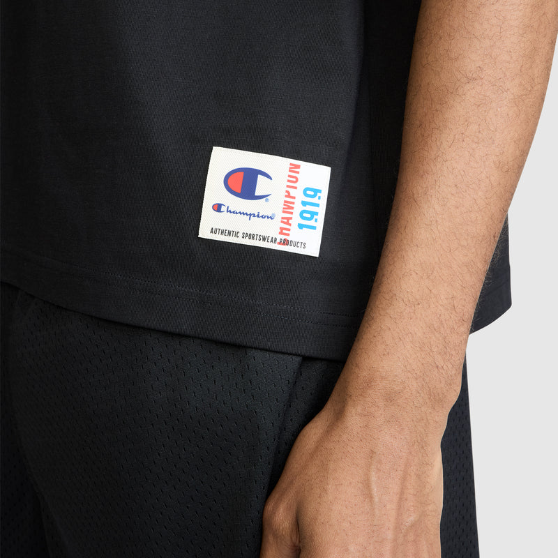 A close-up of a person wearing the Champion Short Sleeve Boxy T-Shirt with Embroidered Script Logo in black and matching shorts, featuring a white label with the Champion logo and text near the hem. This relaxed fit tee showcases the brand's classic heritage style.