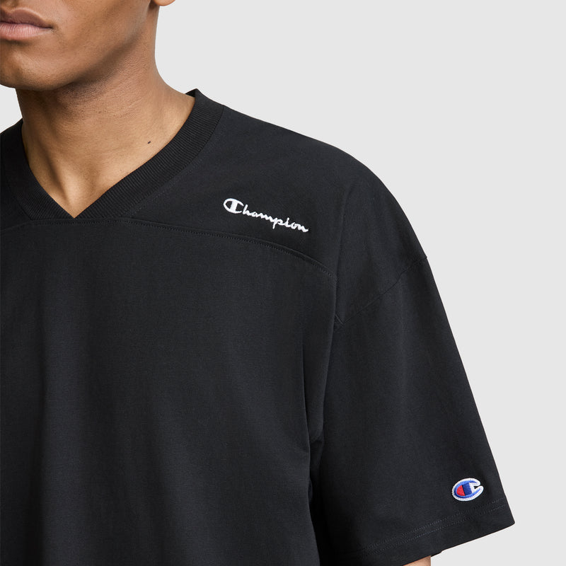 A person is shown from the shoulders up wearing the Champion Short Sleeve Boxy T-Shirt with an embroidered script logo. This relaxed fit men's tee features signature Champion branding on both the chest and sleeve. The plain light gray background emphasizes the heritage style of this classic Champion piece.