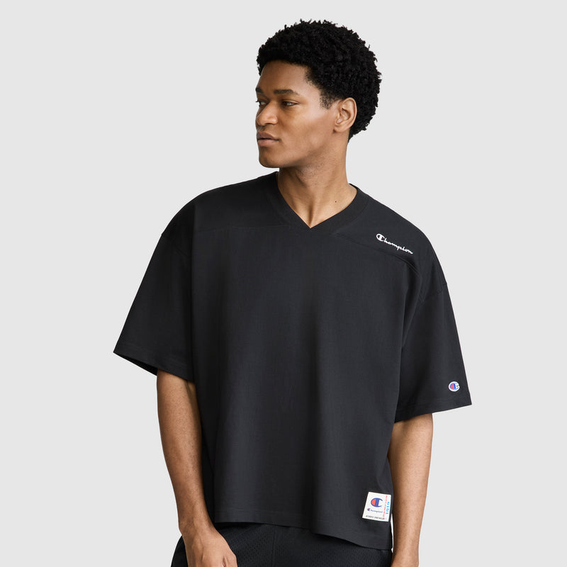 A man wearing a relaxed fit Champion Short Sleeve Boxy T-Shirt with an embroidered script logo—a loose black V-neck tee with short sleeves—stands against a light gray background, looking to his left with a neutral expression.