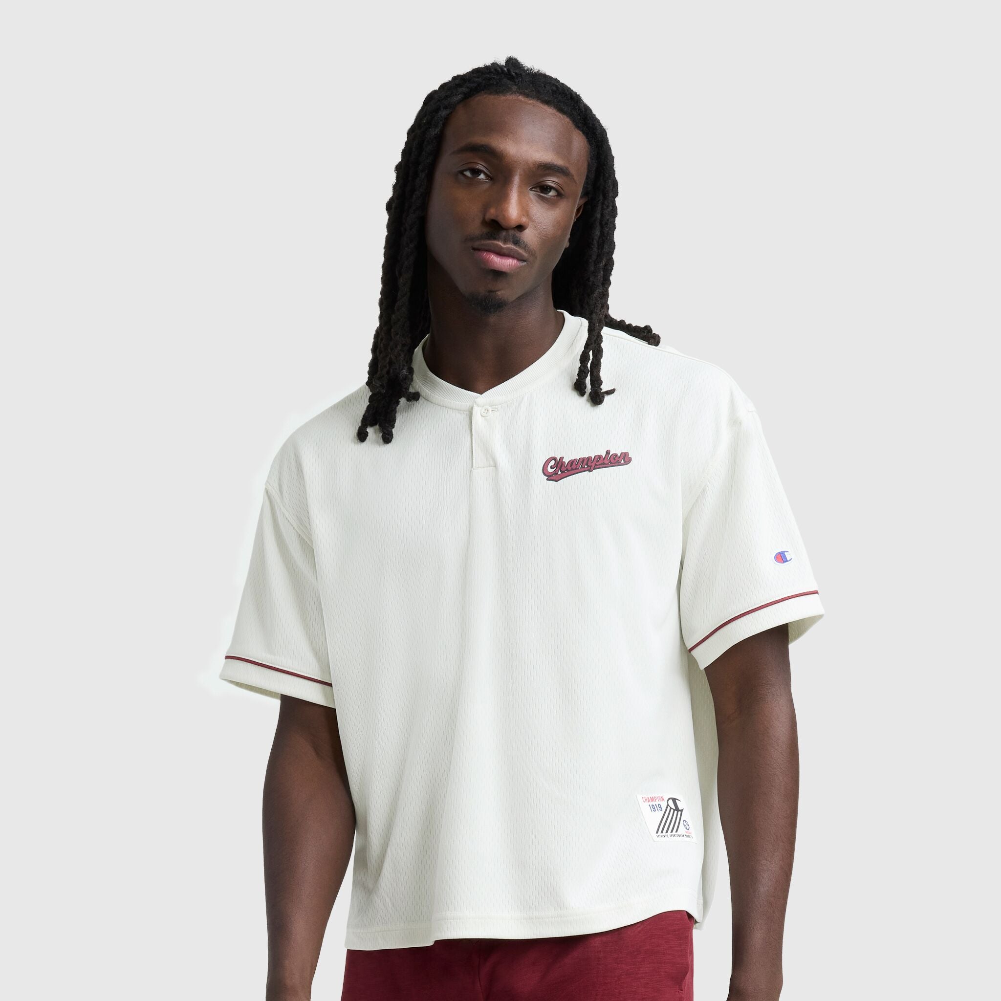 A man with long braided hair wears the Champion Modern Retro Coaches Henley, featuring a C Logo, short sleeves, burgundy trim, and matching shorts for a vintage sport look against a plain light gray background.