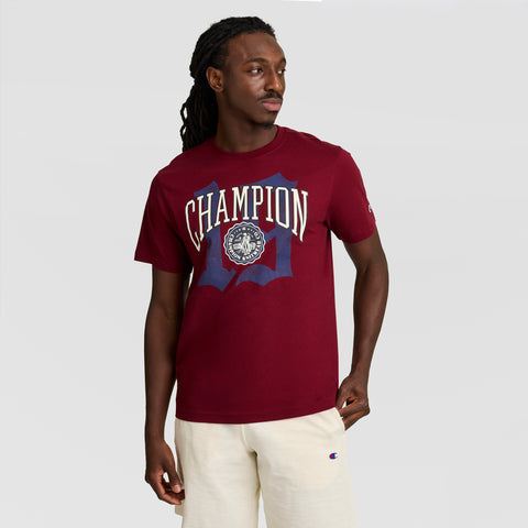 A man in a maroon Champion Modern Retro Gothic '19 T-Shirt with C Logo and light beige shorts stands against a plain background, looking left with one hand in his pocket.
