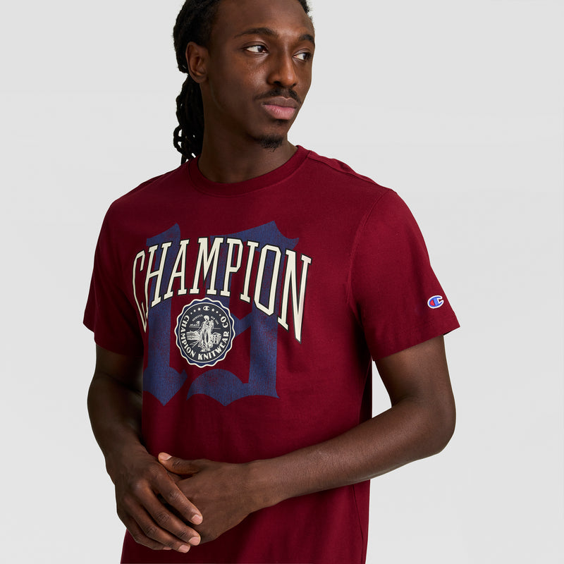 A man with long braided hair wears a maroon Champion Modern Retro Gothic '19 T-Shirt featuring a large C logo graphic on the front. He stands against a plain light gray background, looking to his left.