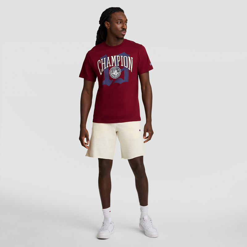 A man stands against a plain gray background wearing a maroon Champion Modern Retro Gothic '19 T-Shirt with C Logo, off-white shorts, white socks, and white sneakers. He looks to his left with his hands relaxed by his sides.