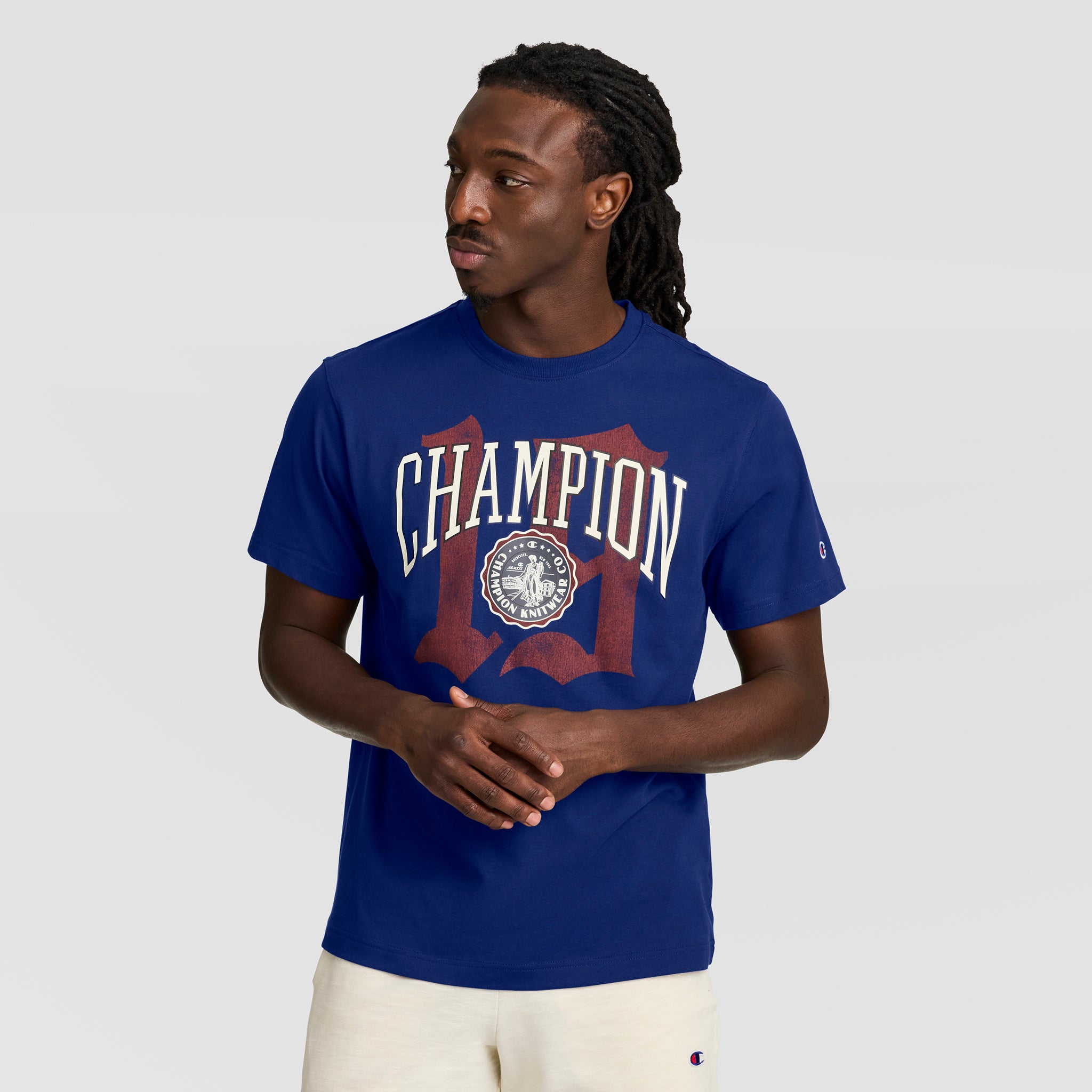 A man with long braided hair wears a blue Champion Modern Retro Gothic '19 T-Shirt with the C logo and light-colored shorts. He stands against a plain gray background, looking to the side with hands loosely clasped in modern retro style.