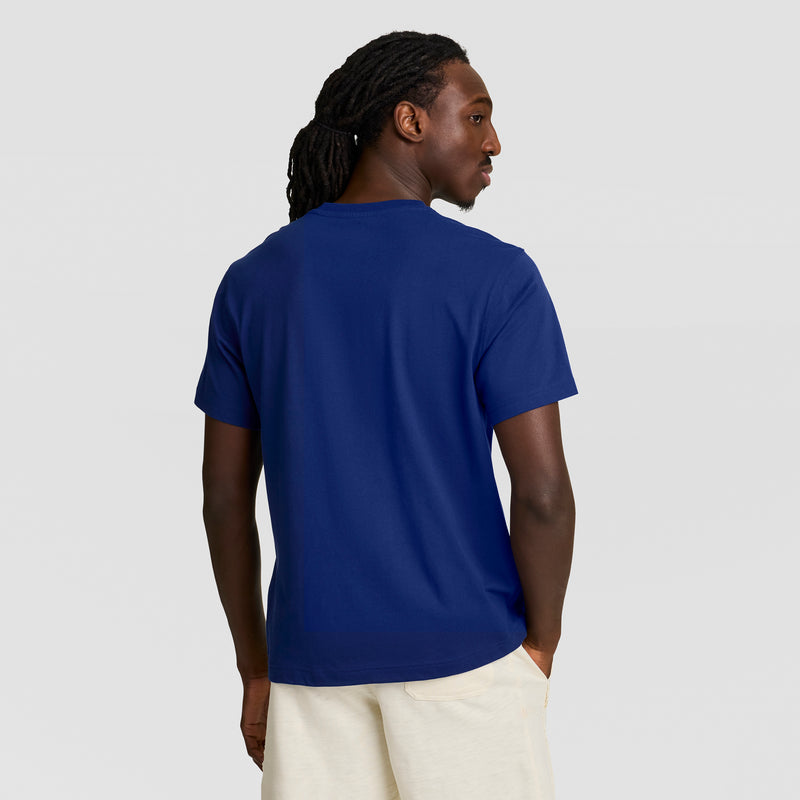 A person with long, dark dreadlocks stands with their back to the camera, showcasing a Champion Modern Retro Gothic '19 T-Shirt with C Logo and light-colored shorts against a plain light gray background.