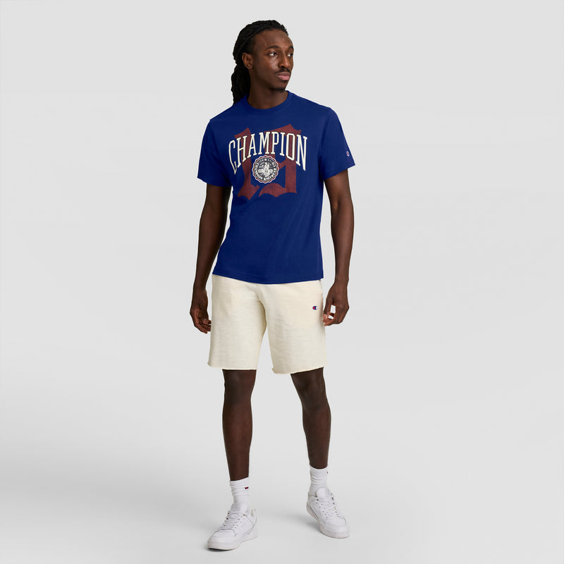 A man in a blue Champion Modern Retro Gothic '19 T-Shirt with C logo, light beige shorts, white socks, and white sneakers stands against a plain light gray background, looking slightly left—a clean modern retro style.