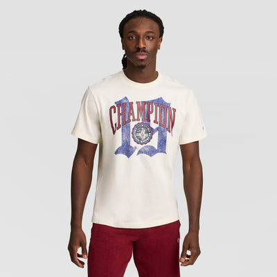 A man with long braided hair wears a cream Modern Retro Gothic '19 T-Shirt, C Logo by Champion in red and blue, paired with red pants. He stands against a plain light gray background.